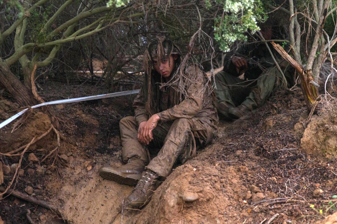 A Marine sits in the dirt wearing a camouflage suit.