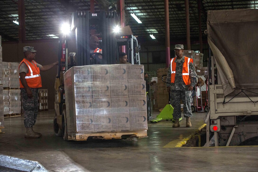 A forklift lifts a pallet of boxes with two service members observing nearby.