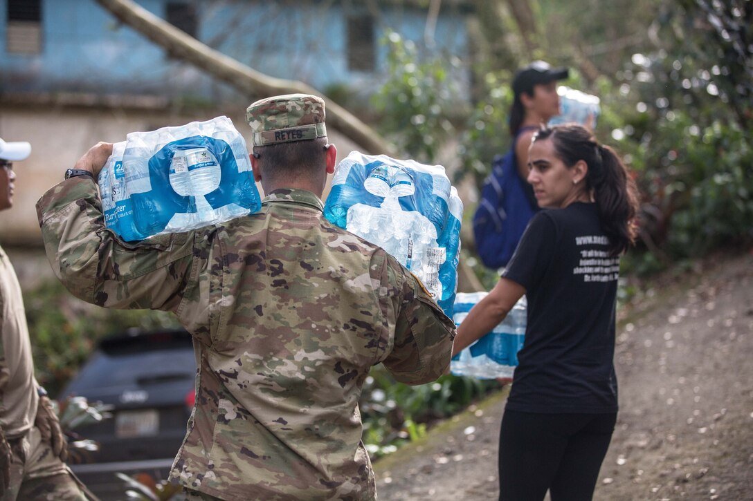 A soldier and civilians carry cases of bottled water.