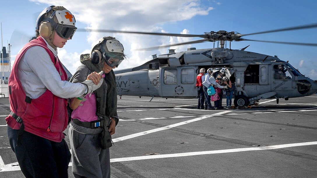 A sailor assists a person walking while other sailors help people out of a helicopter.