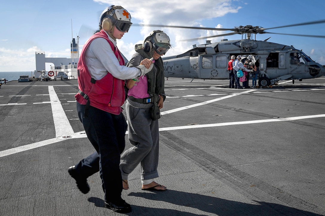 A sailor assists a person walking while other sailors help people out of a helicopter.