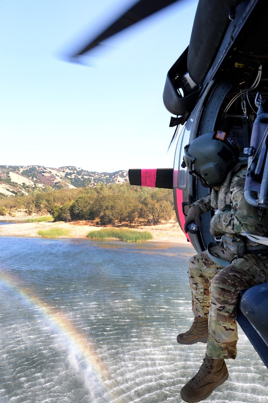 A guardsman sits out the open door of a flying helicopter above water.