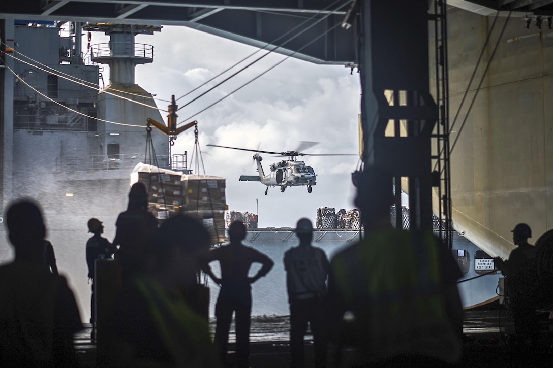 Sailors on one ship watch a helicopter hover over another ship.