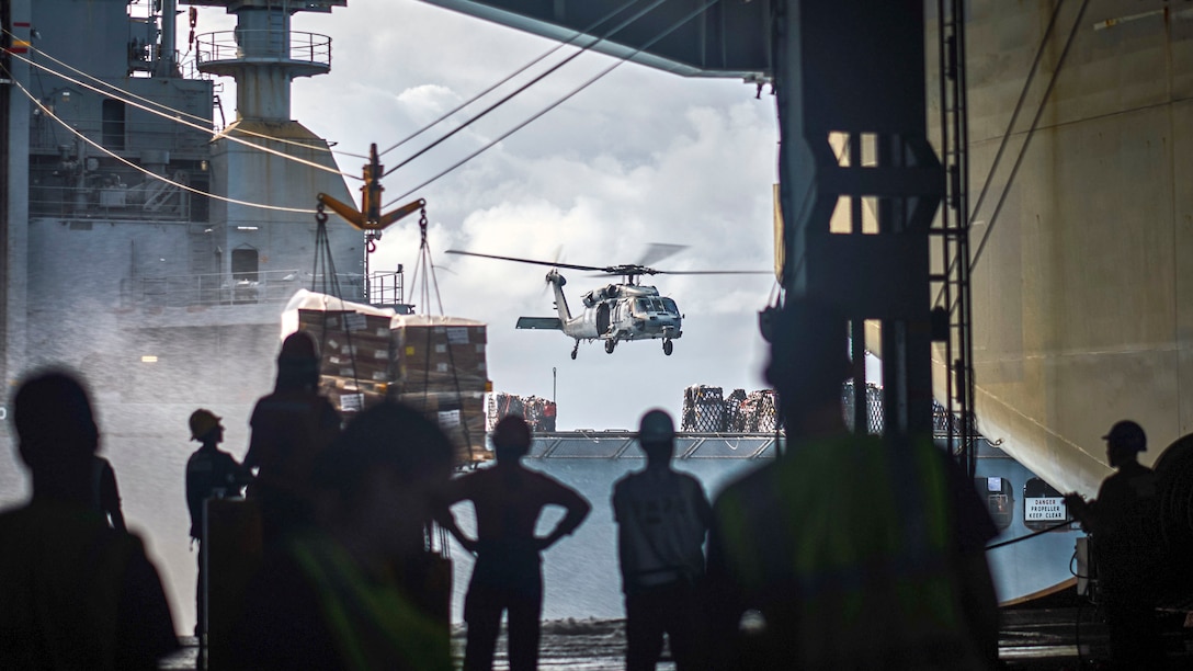 Sailors on one ship watch a helicopter hover over another ship.