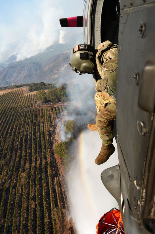 A soldier leans out the open door of a flying helicopter while dropping water.