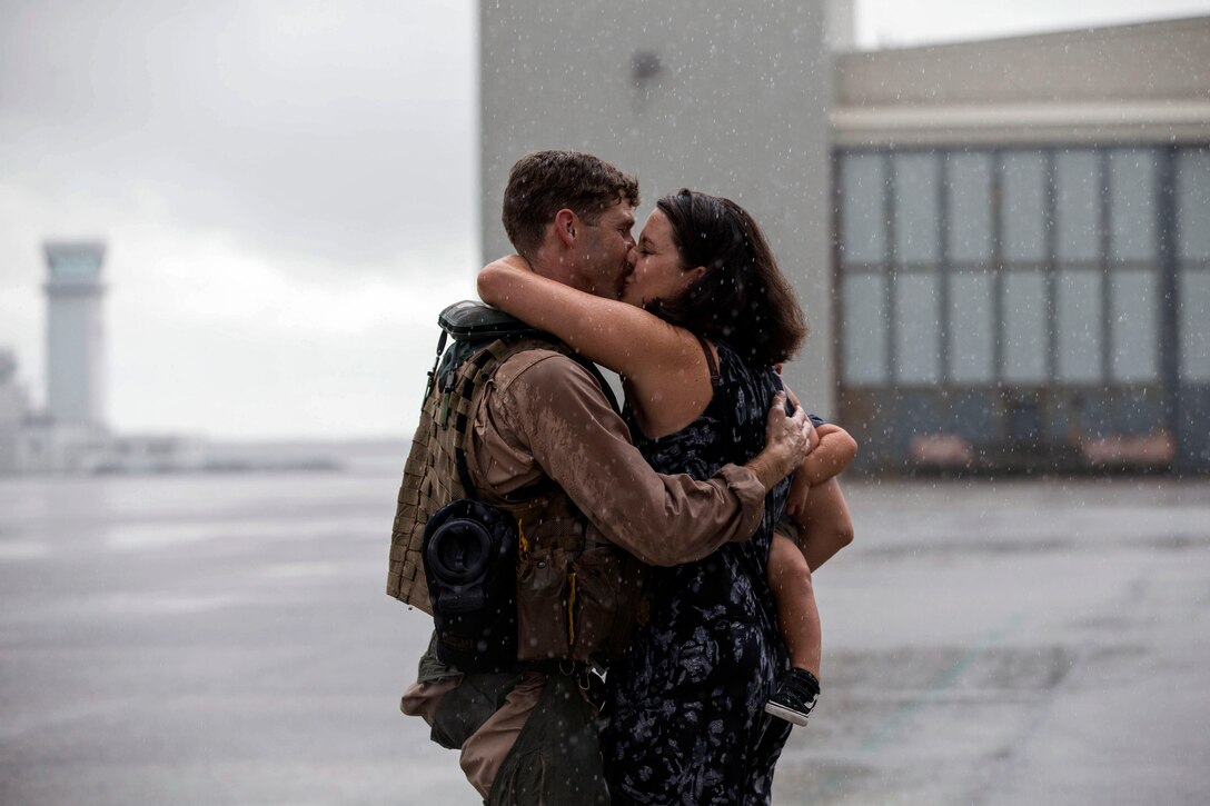 A man kisses a woman holding a baby on a flightline.