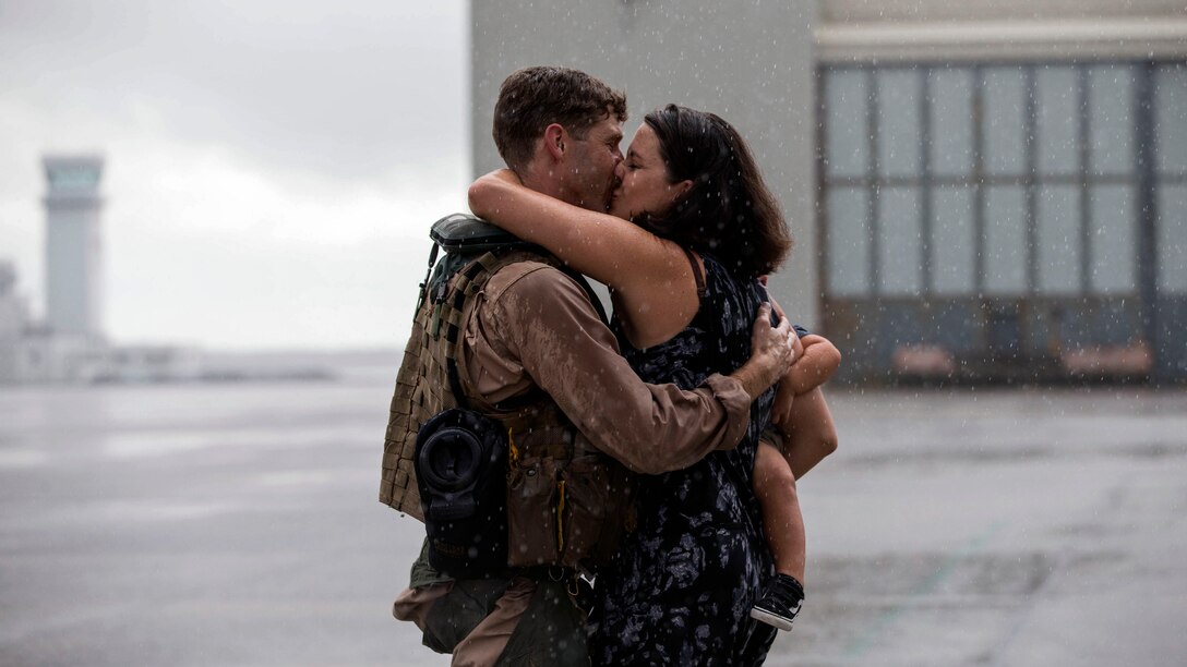 A man kisses a woman holding a baby on a flightline.