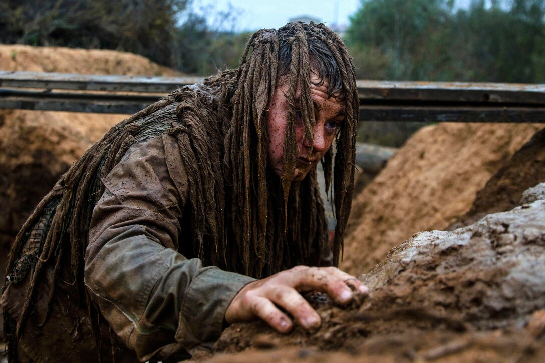 A Marine in camouflage crawls out of a mud pit.