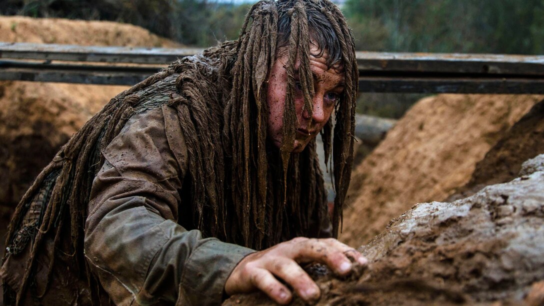 A Marine in camouflage crawls out of a mud pit.