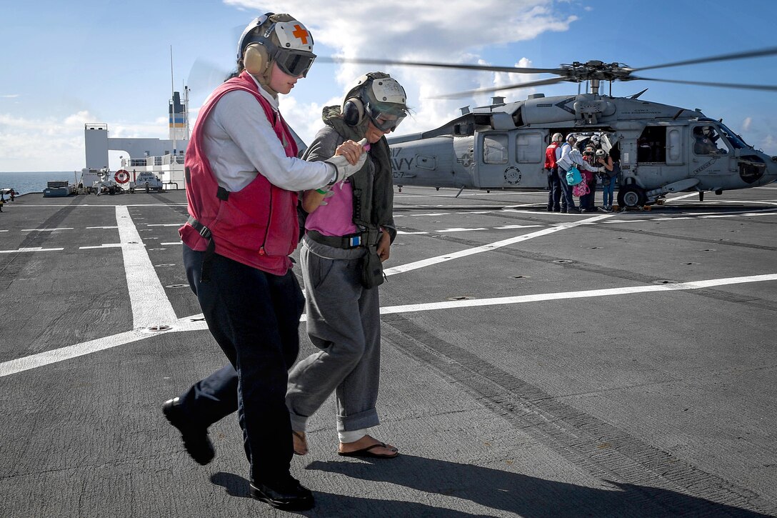 A sailor assists a person walking while other sailors help people out of a helicopter.