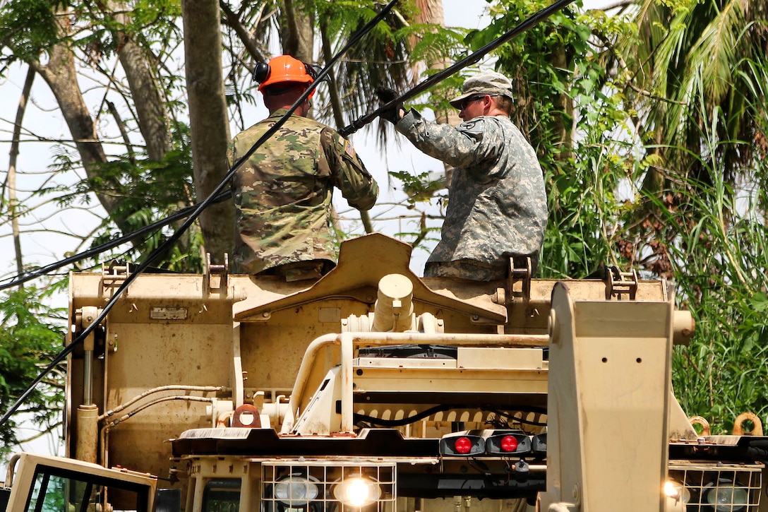 Two guardsmen sit on machinery and clear branches from power lines.