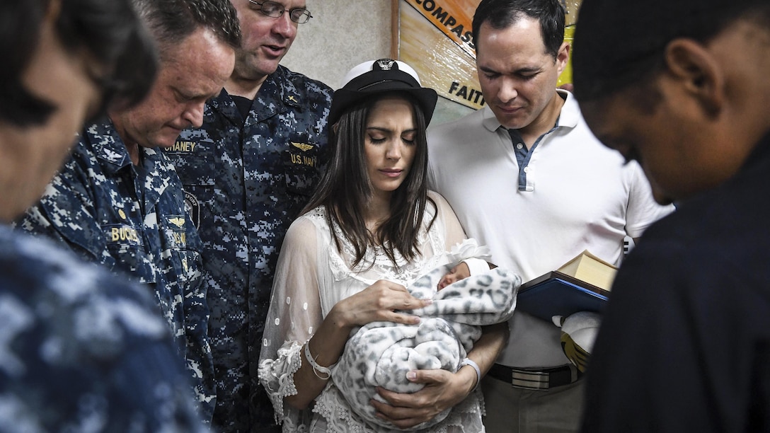 A woman holding a baby is surrounded by people with their heads lowered.