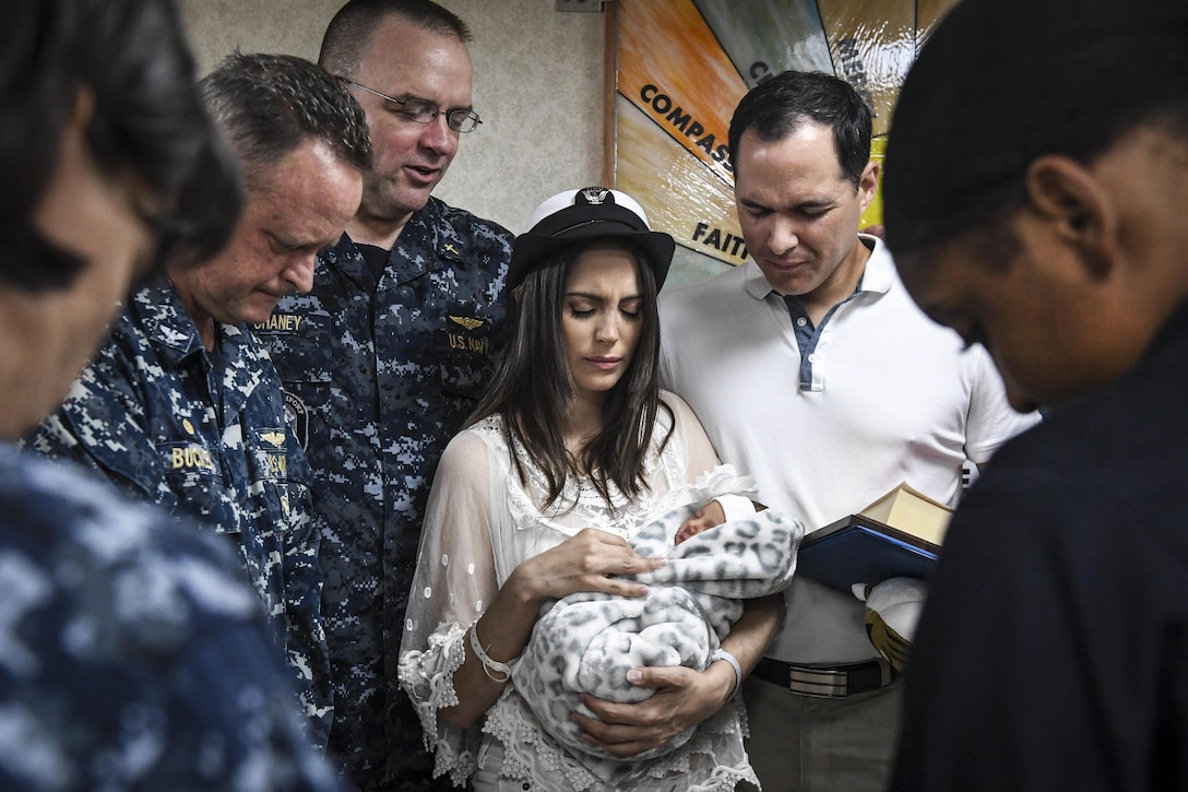 A woman holding a baby is surrounded by people with their heads lowered.