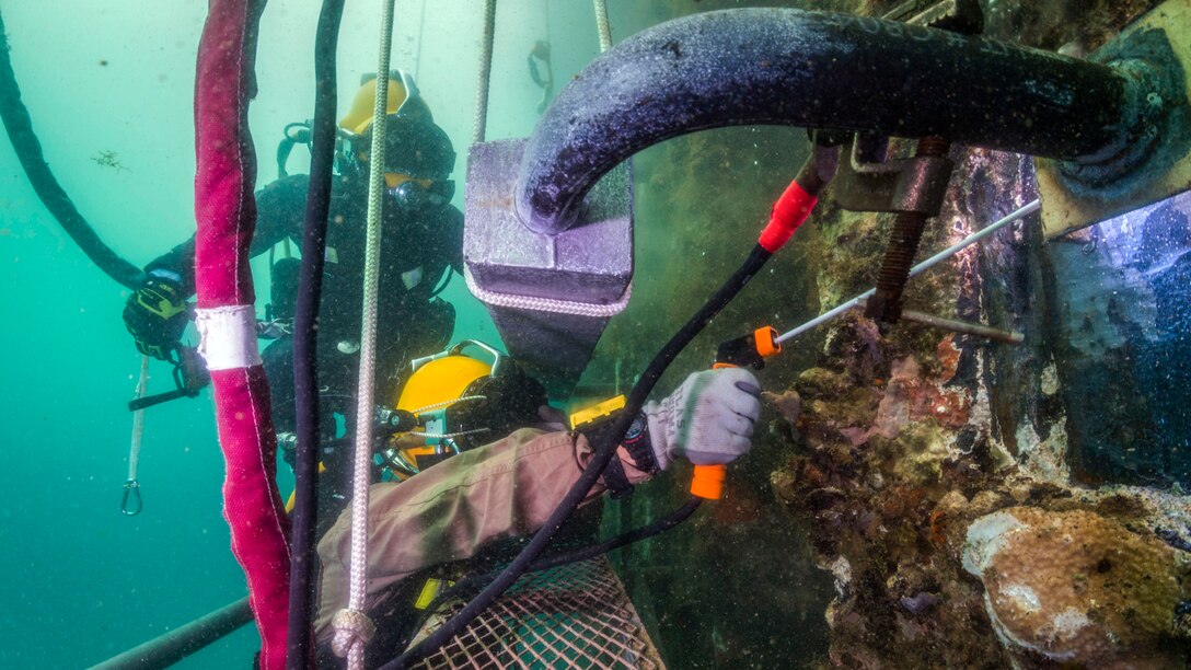 Two sailors in diving gear perform welding work on underwater structures.