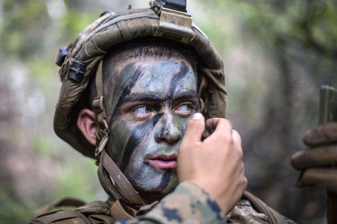 A Marine looks in a compact mirror while putting on camouflage paint in wooded surroundings.