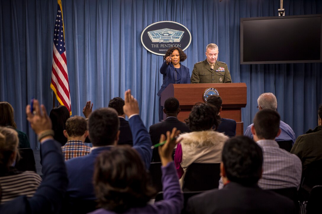 Two people standing at a lectern take questions from seated reporters raising their hands.