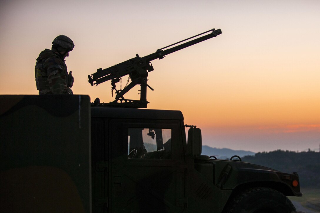 An airman, shown in silhouette, sits atop a vehicle and mans a machine gun.