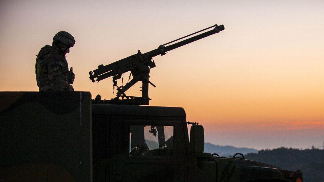 An airman, shown in silhouette, sits atop a vehicle and mans a machine gun.