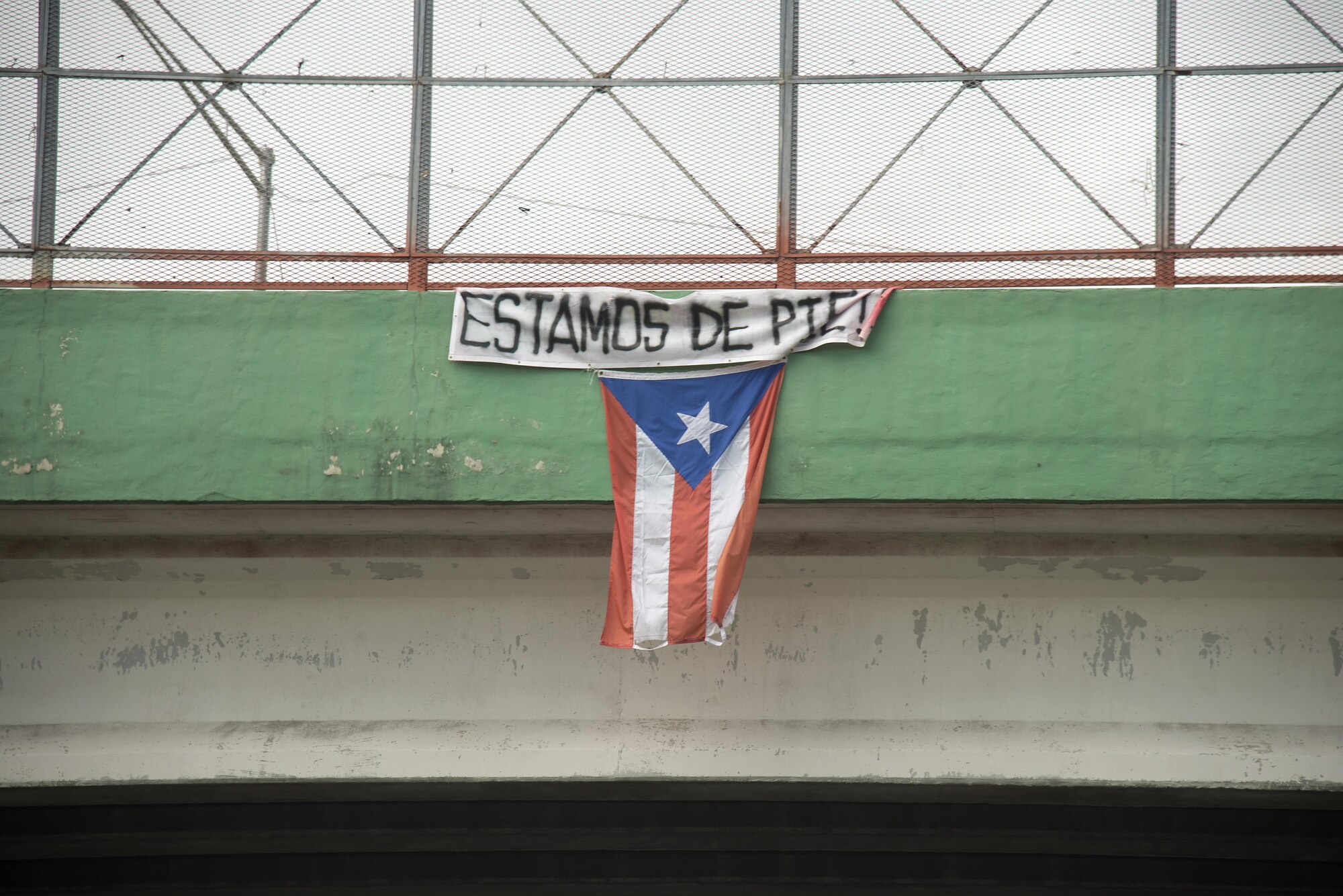 A flag hangs from a highway overpass that reads “Estamos de pie,” in Caguas, Puerto Rico, Oct. 2, 2017. “Estamos de pie” translates to “We are standing,” which is meant to motivate people to stay strong and overcome the disaster left behind Hurricane Maria.