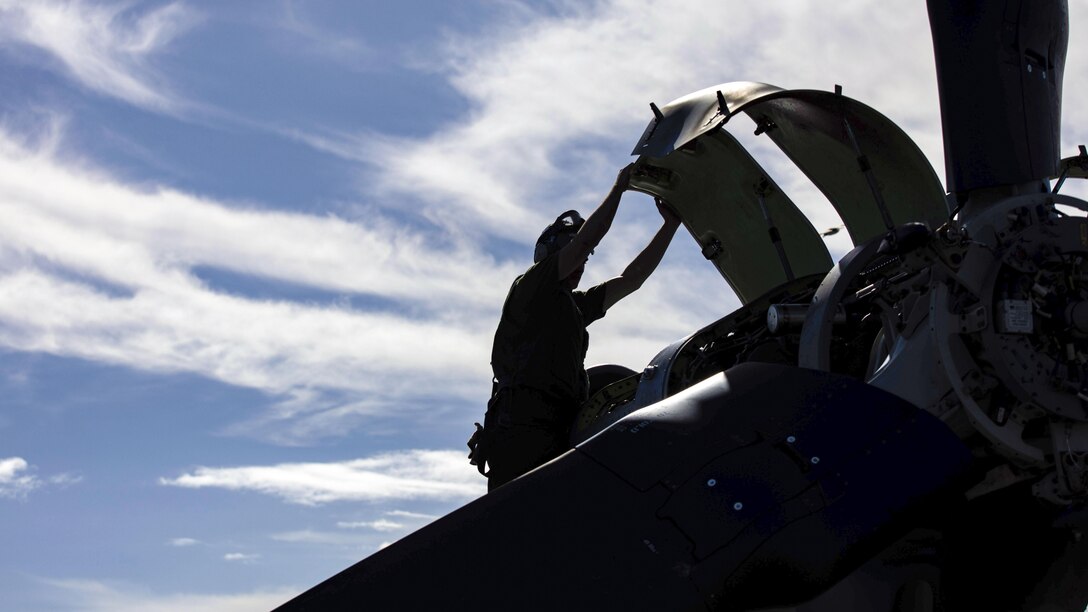 A Marine, shown in silhouette, reaches up and holds part of an aircraft's external structure.