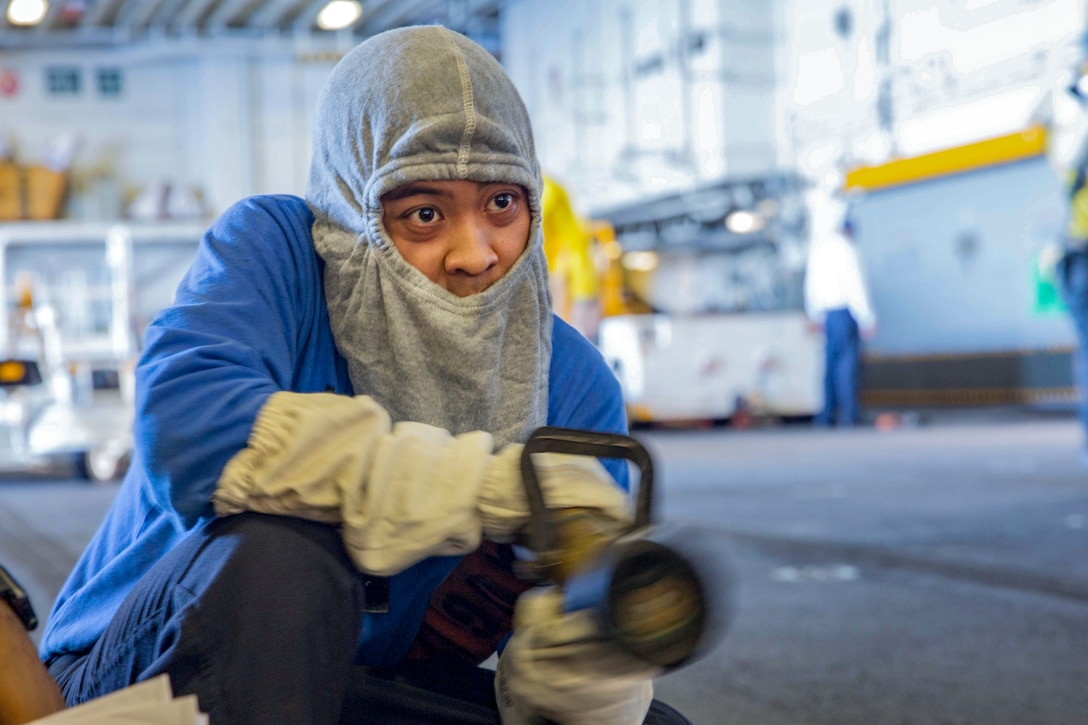 A sailor in a blue shirt and gray balaclava wields a hose in a ship's hangar bay.