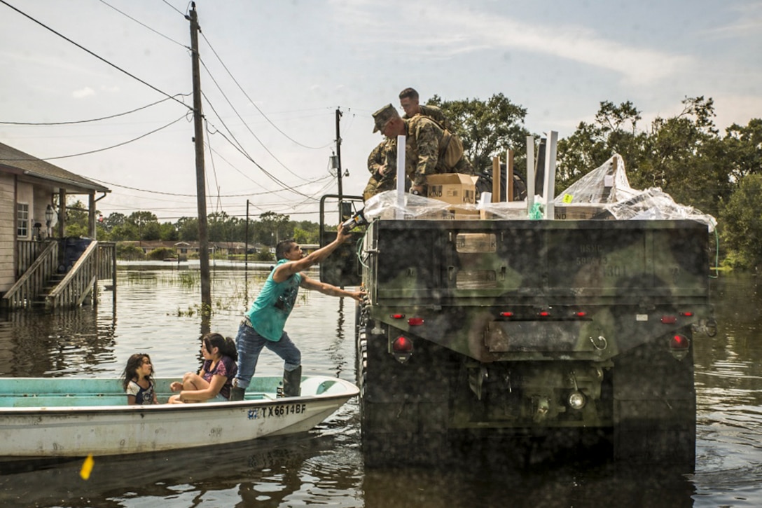 Marines in a military vehicle in a flooded area hand supplies to a man in a boat with two girls.