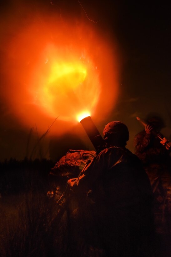 A soldier fires a mortar system at night, showing a fireball.