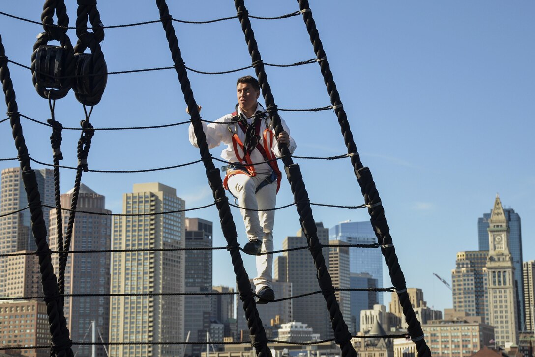 A sailor climbs the mizzenmast on a ship with the Boston skyline in the background.