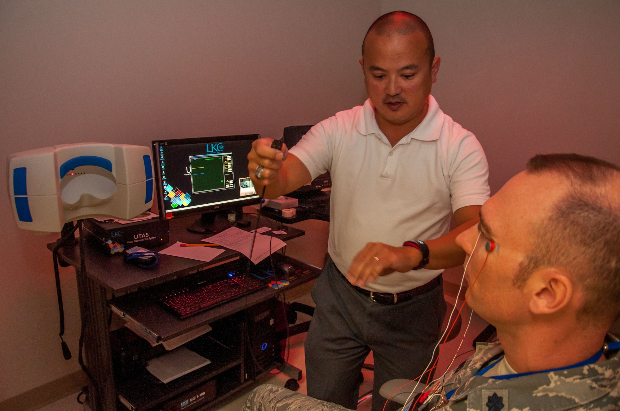 Dino Tsuchiyama (left), Aeromedical Consultation Service Ophthalmology Branch electro-diagnostic technician, attaches electrodes from Lt. Col. Christopher Cannon to a multifocal electroretinogram machine. The machine measures electrical activity in the retina when the eye is exposed to a light source. (U.S. Air Force photo/John Harrington)