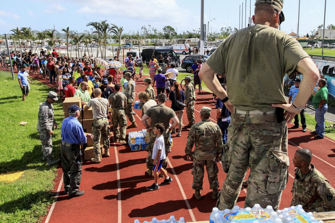 An Army commander stands on a truck as soldiers prepare to deliver food and water.