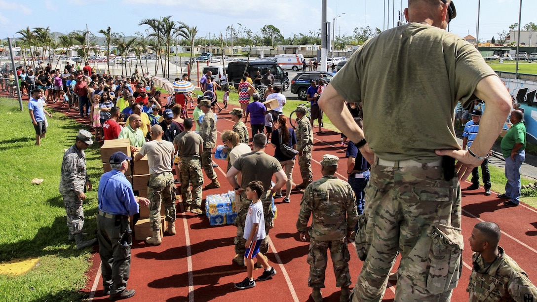 An Army commander stands on a truck as soldiers prepare to deliver food and water.