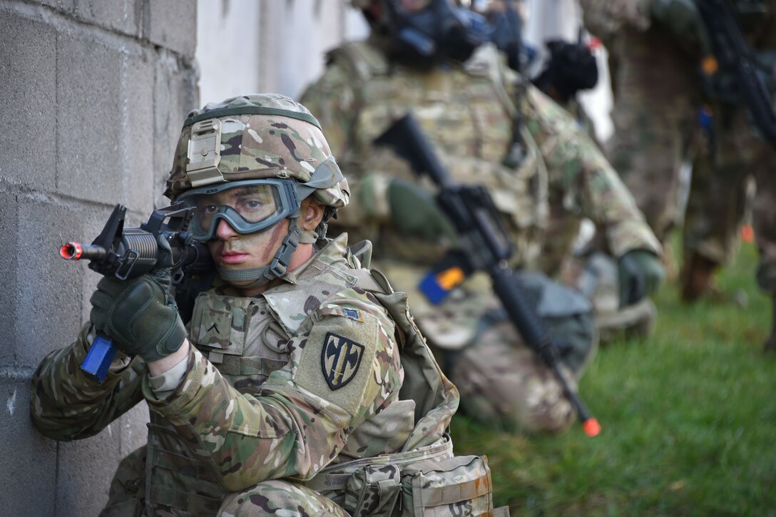 A soldier with a paintball rifle kneels next to a building.