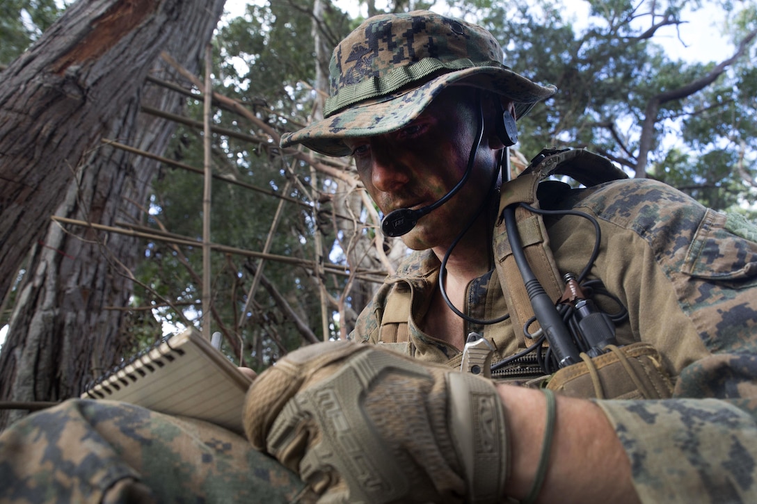 A Marine with a camouflaged face and wearing a boonie cap takes notes during a jungle patrol.