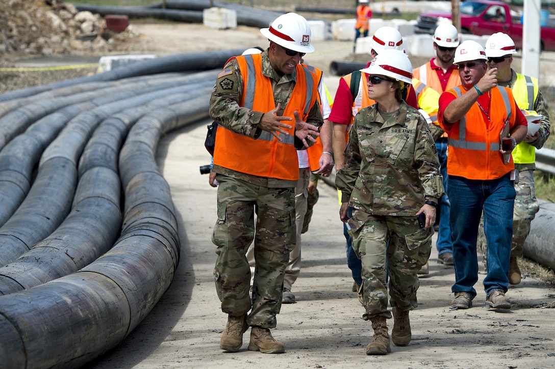 Service members and civilians in hard hats converse while walking past snaking black pipes.