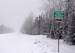 A sign welcoming visitors to Clear Air Force Station stands next to a small road covered in snow.