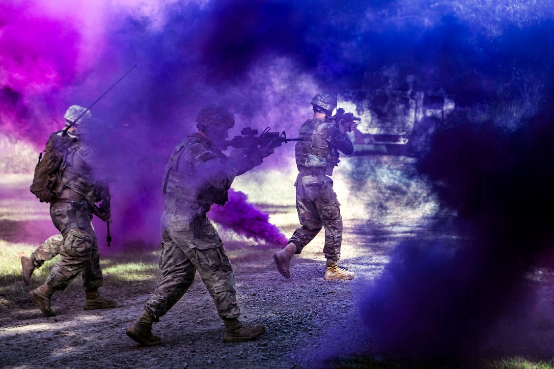 Airmen walk and point weapons as purple smoke wafts around them.