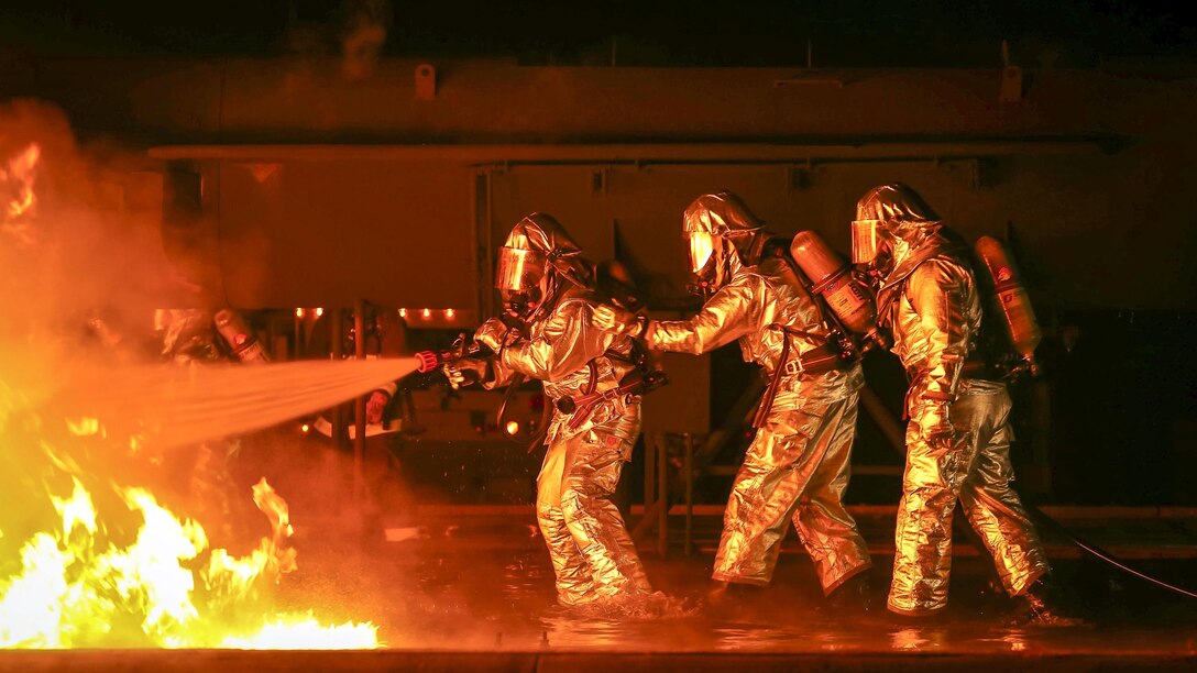 Three Marines in silver protective gear spray water from a hose onto a blaze.