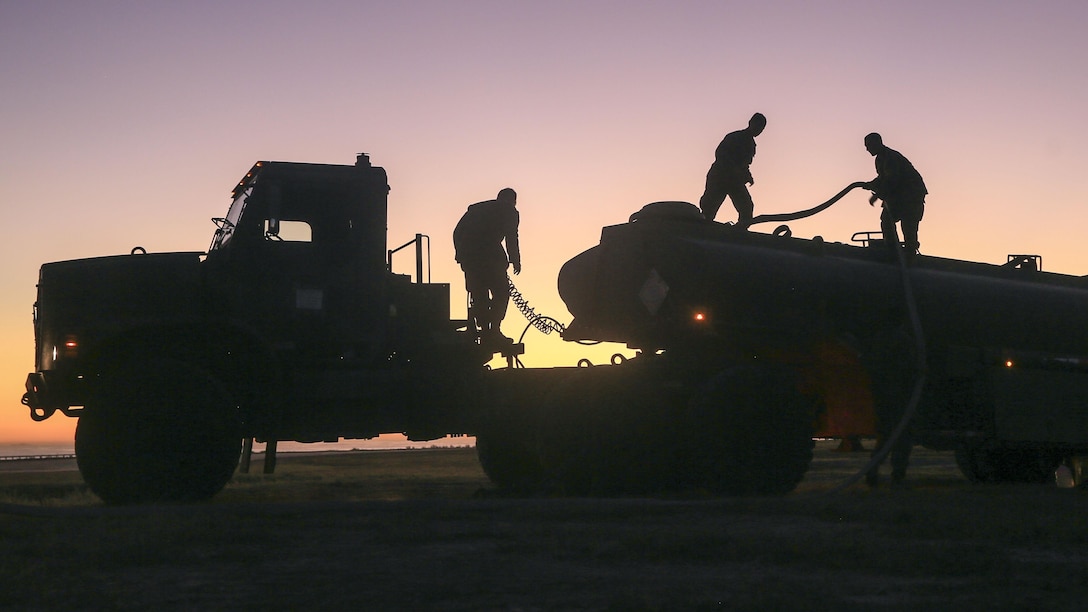 Marines, shown in silhouette, work with hoses atop a semitrailer against a light purple sky.