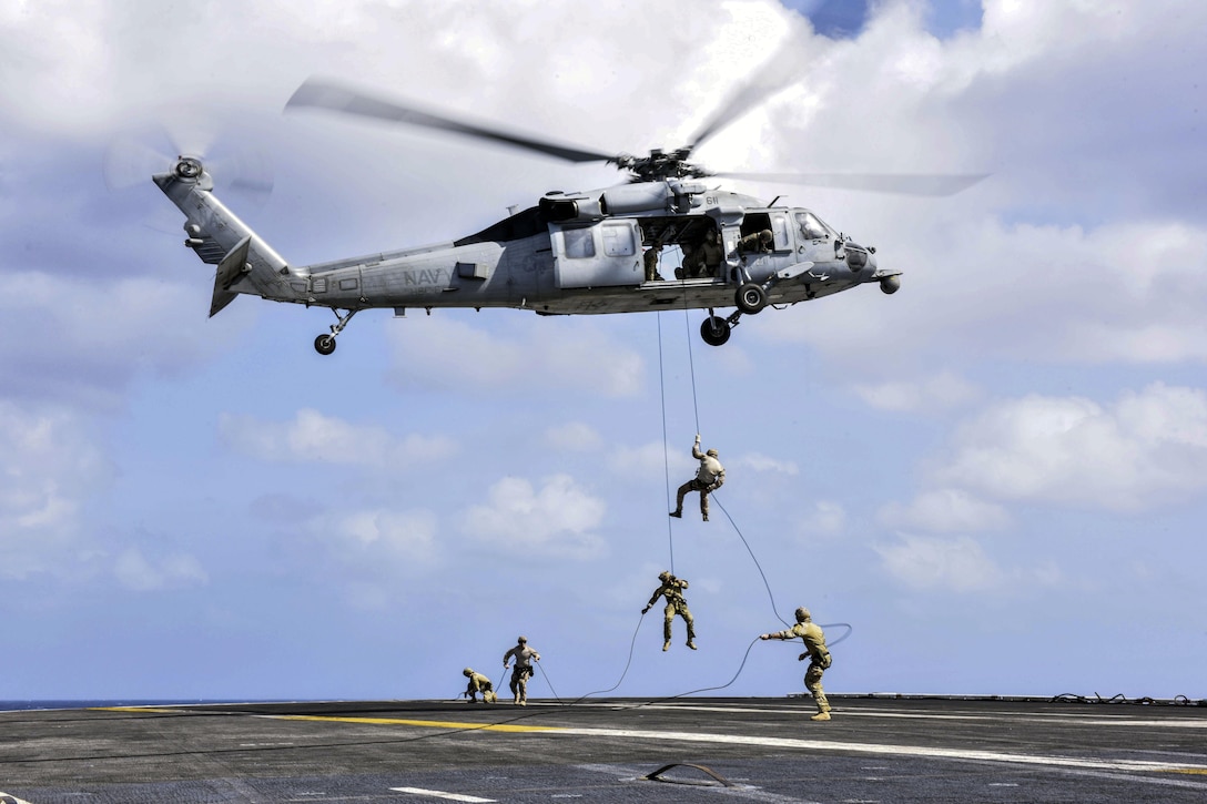 Sailors rappel from a helicopter onto a ship's flight deck.