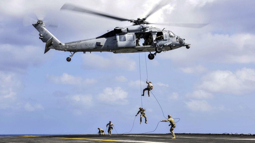 Sailors rappel from a helicopter onto a ship's flight deck.
