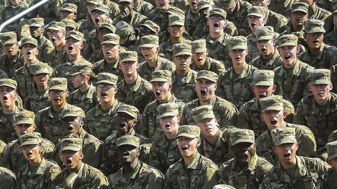 Soldiers gathered on bleachers open their mouths wide while performing a call.