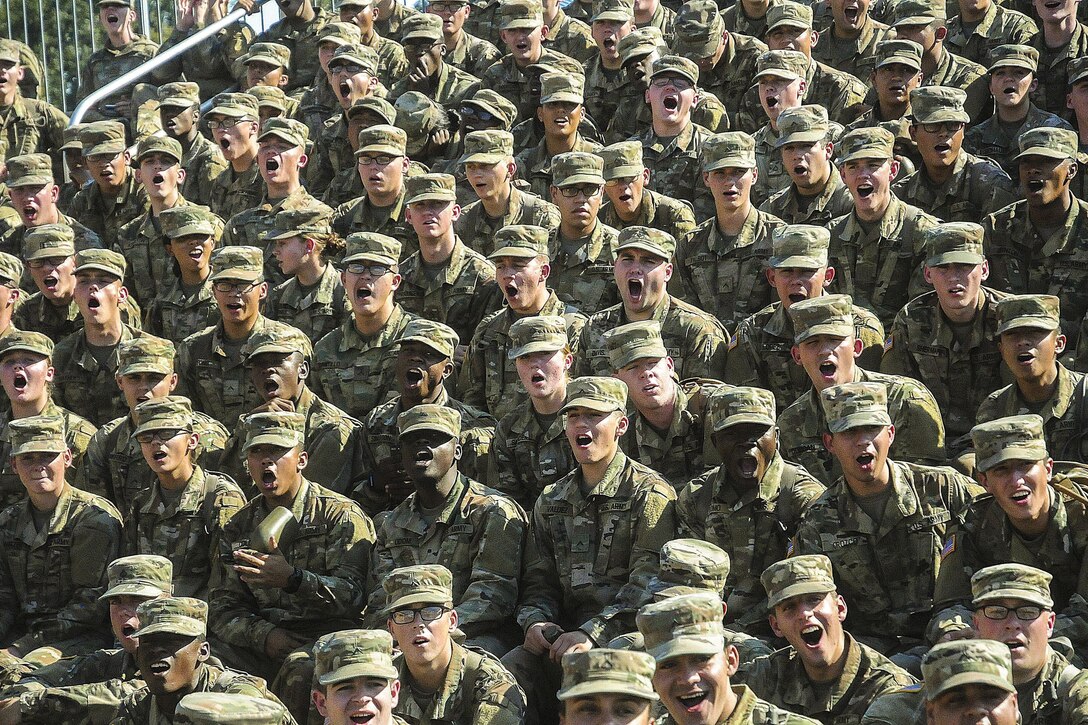 Soldiers gathered on bleachers open their mouths wide while performing a call.