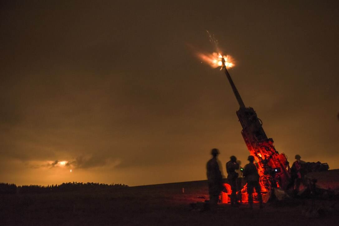Soldiers stand around a howitzer as it fires at night, creating a red glow around the troops.