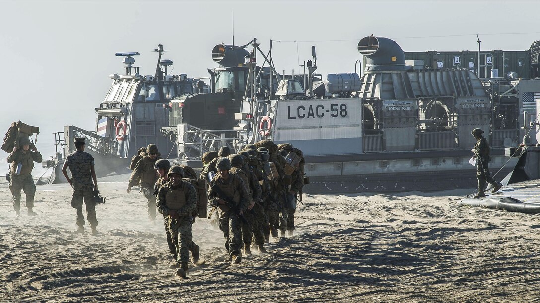 A group of Marines runs on a beach away from a landing craft parked in the sand.