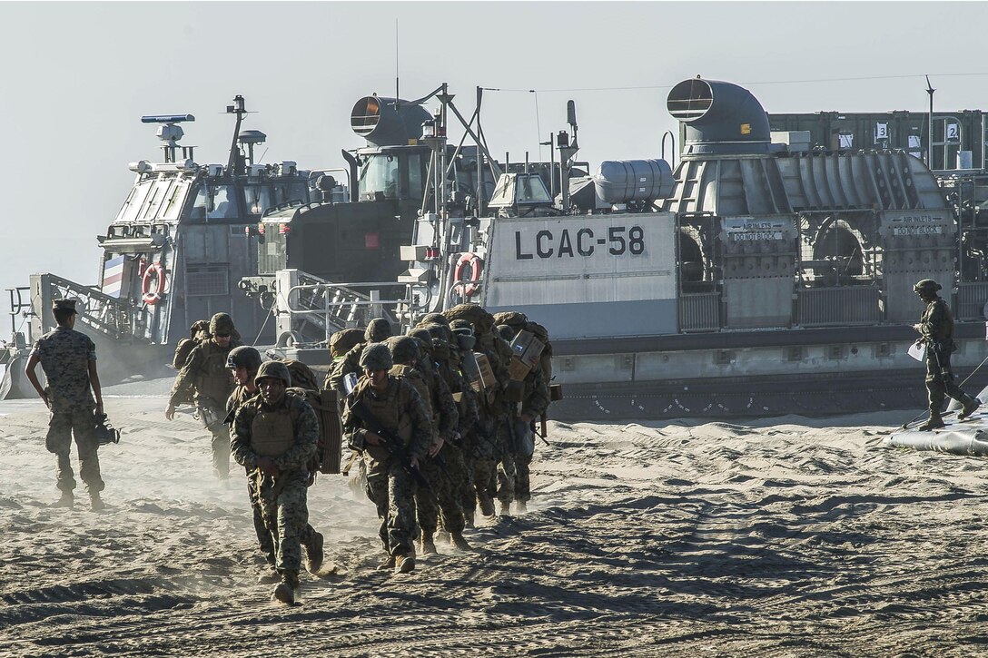 A group of Marines runs on a beach away from a landing craft parked in the sand.