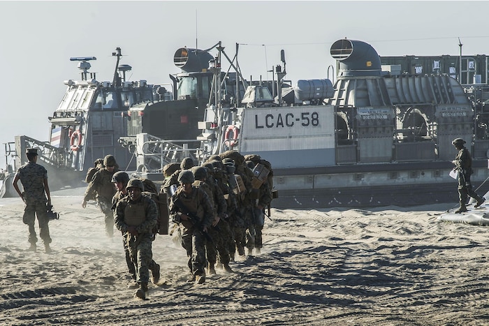 A group of Marines runs on a beach away from a landing craft parked in the sand.