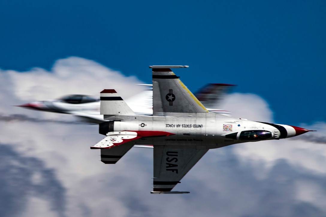 Two fighter jets cross in the sky, one flying straight ahead and the other on its side.