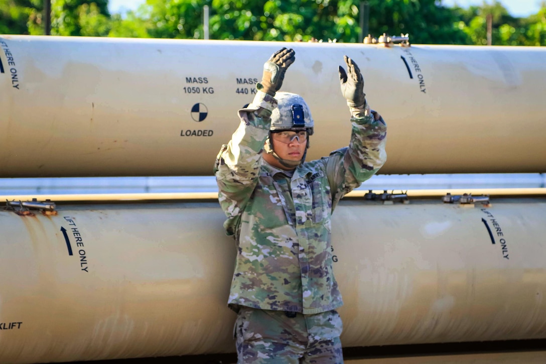 A soldier uses hand signals to guide a military forklift to lift a missile round trainer.