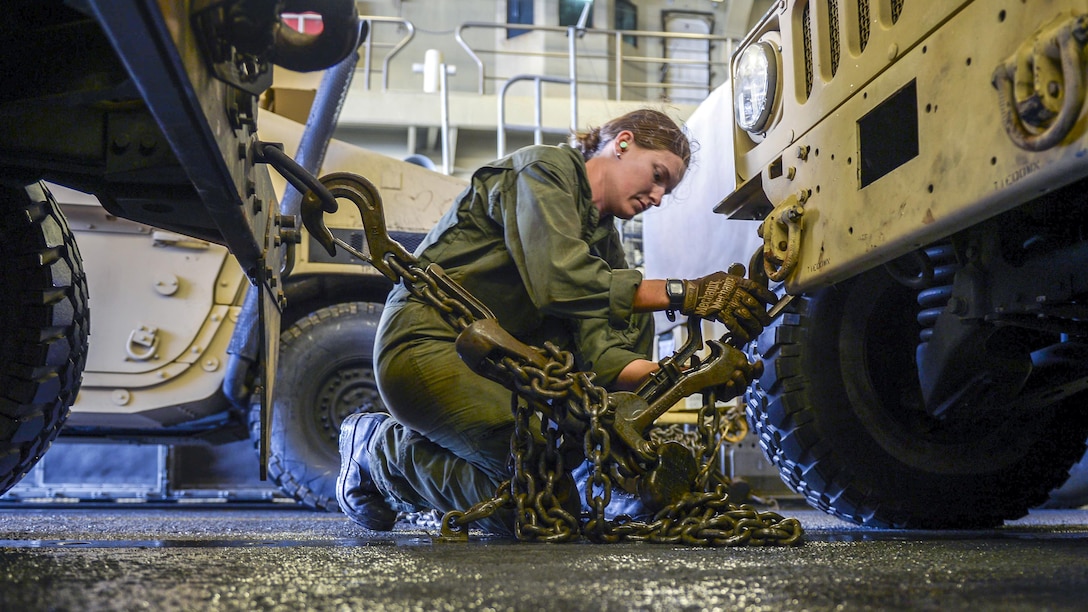 A sailor kneels and chains the front of a vehicle to a metal loop on the floor.