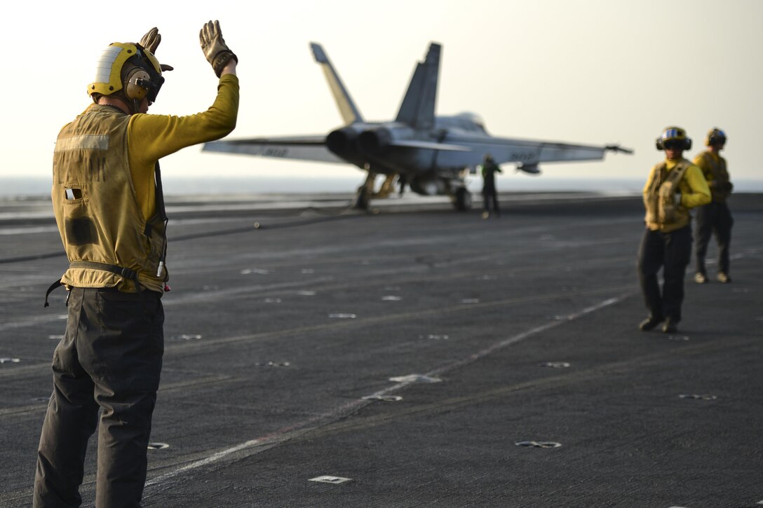 A sailor gives a hand signal with an aircraft in the background.